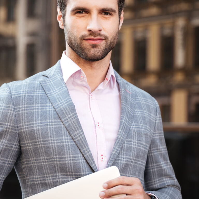 Portrait of a young handsome man in jacket holding laptop computer and looking at camera while standing in a city area